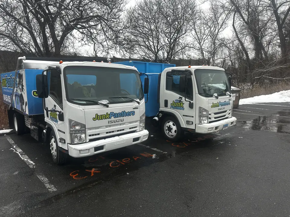 Two white Junk Panthers Isuzu trucks with blue dump bodies parked in a lot after a snowfall